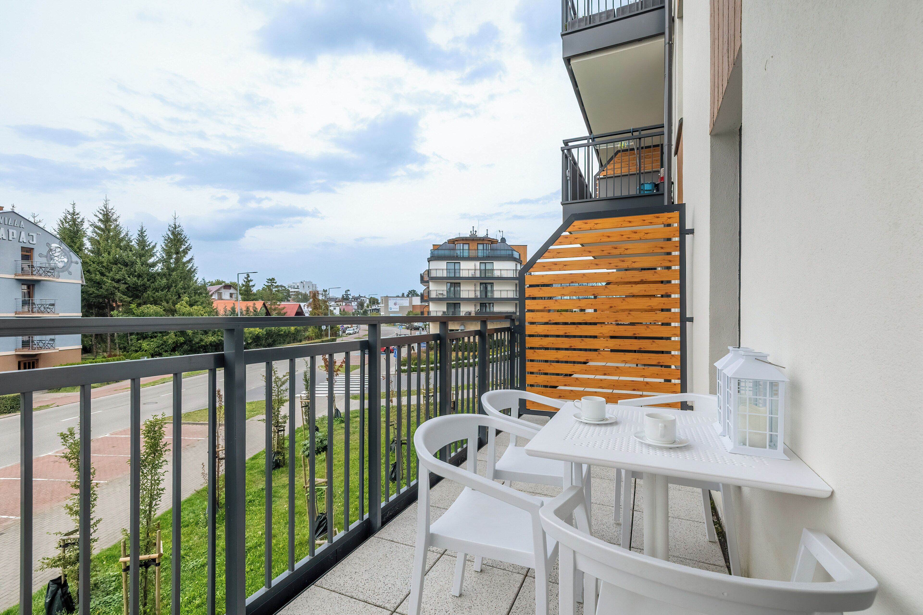Ein Balkon mit weißen Stühlen und einem Tisch, mit Blick auf eine Straße und Grünflächen.