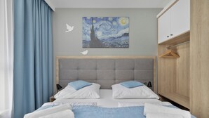 Een net opgemaakt bed met wit- en blauw beddengoed, een grijze hoofdbord en een schilderij van een nachthemel aan de muur.
