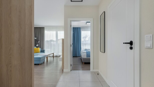 Een gang die leidt naar een slaapkamer met een bed, nachtkastje en een raam met gordijnen.