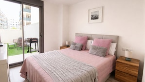 Um quarto com cama, mesas de cabeceira, uma mesinha de apoio, uma janela com vista para edifícios e uma porta que dá para uma varanda.