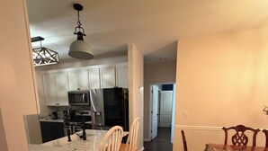 Una cocina con armarios blancos, microondas y refrigerador. Hay un comedor con mesa y sillas.