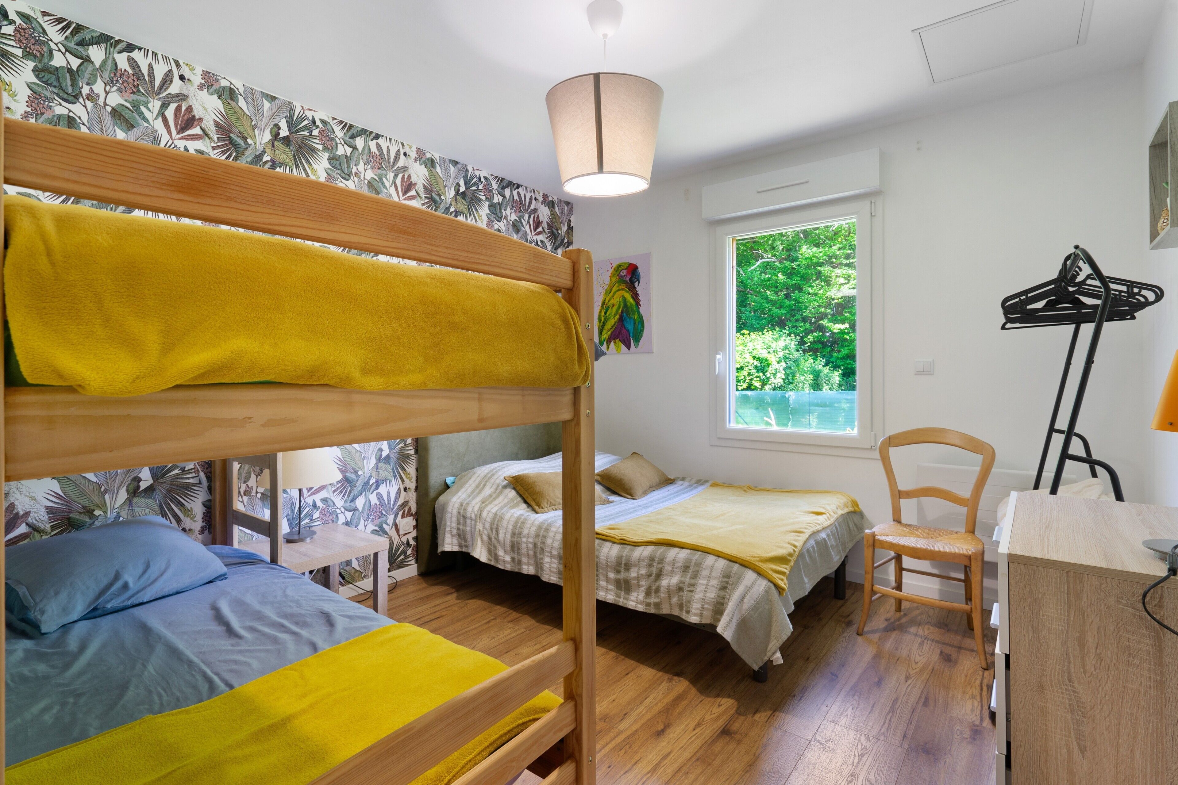 A dormitory room with bunk beds, a desk, a chair, and a window with a view of greenery.