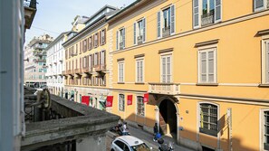 A yellow building with multiple windows and balconies, a street with cars and a motorcycle, and a pedestrian walkway. - Italianway Comfort - Dell'Orso 9 A (Milan)