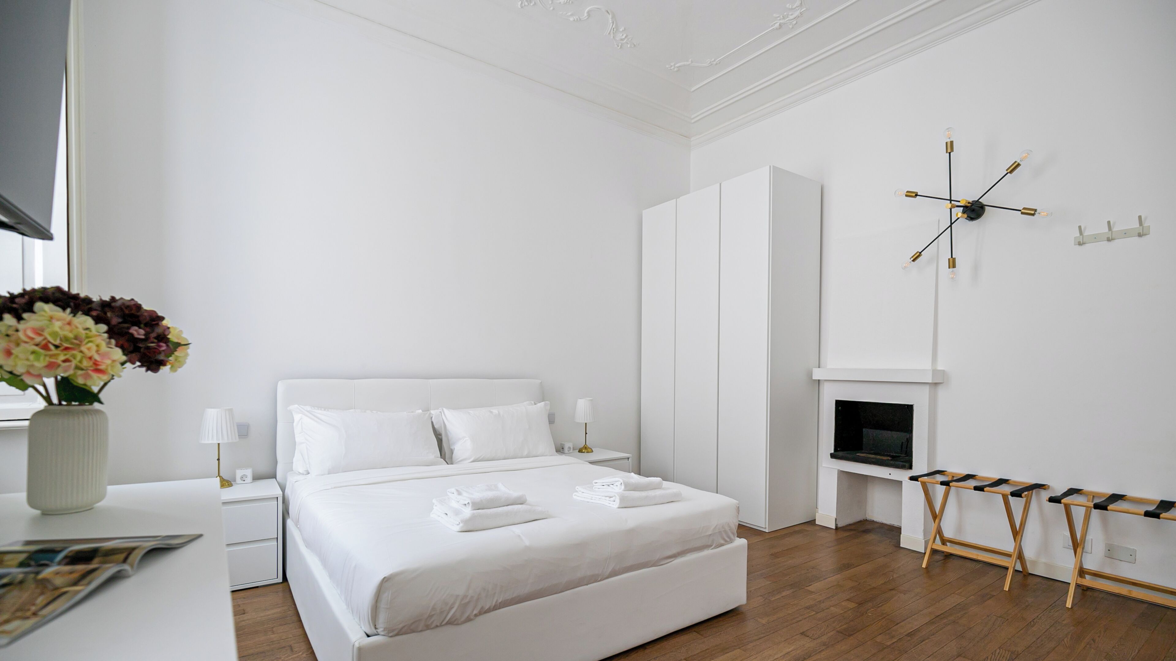 Une chambre moderne avec un grand lit, des murs blancs, un plancher en bois, une armoire blanche, une petite table avec une lampe et une horloge murale.