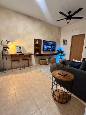 A living room with a TV, a ceiling fan, a sofa, a coffee table, and a dining area with chairs and a lamp. - Casa Oasis #4 - Cozy 1br apartment in lovely San Juan with AC and Solar Power (San Juan)