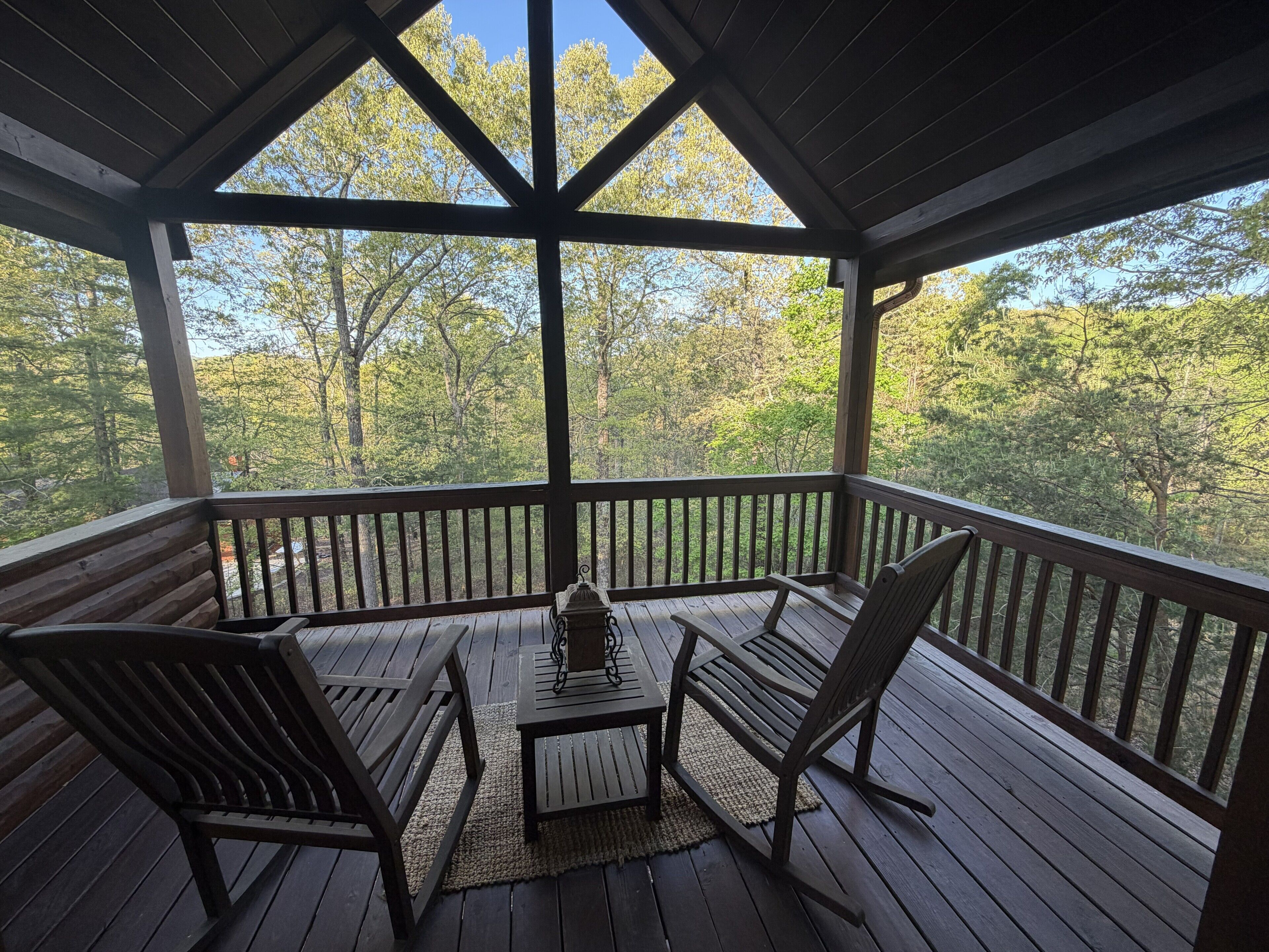 A covered outdoor deck with wooden furniture, a table, and a view of a forested area.