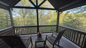 A covered outdoor deck with wooden furniture, a table, and a view of a forested area. - Tranquility Cabin: Secluded Mountain Escape with Game Room, Fire Pit & Hot Tub (Blue Ridge)