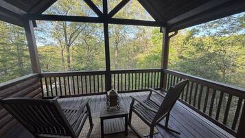 A covered outdoor deck with wooden furniture, a table, and a view of a forested area.