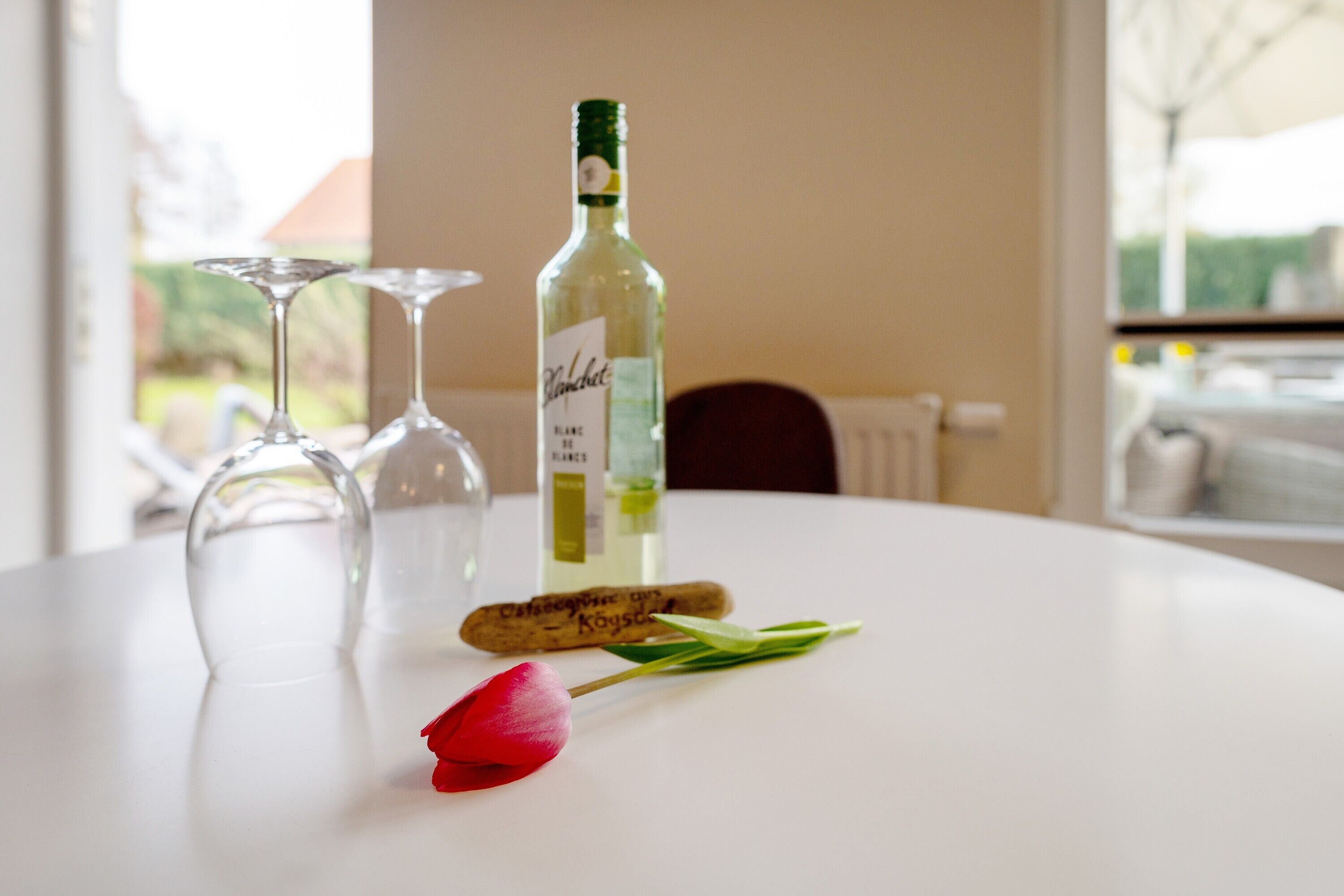 Eine Weinflasche, zwei leere Weingläser und eine rote Tulpe auf einem weißen Tisch.