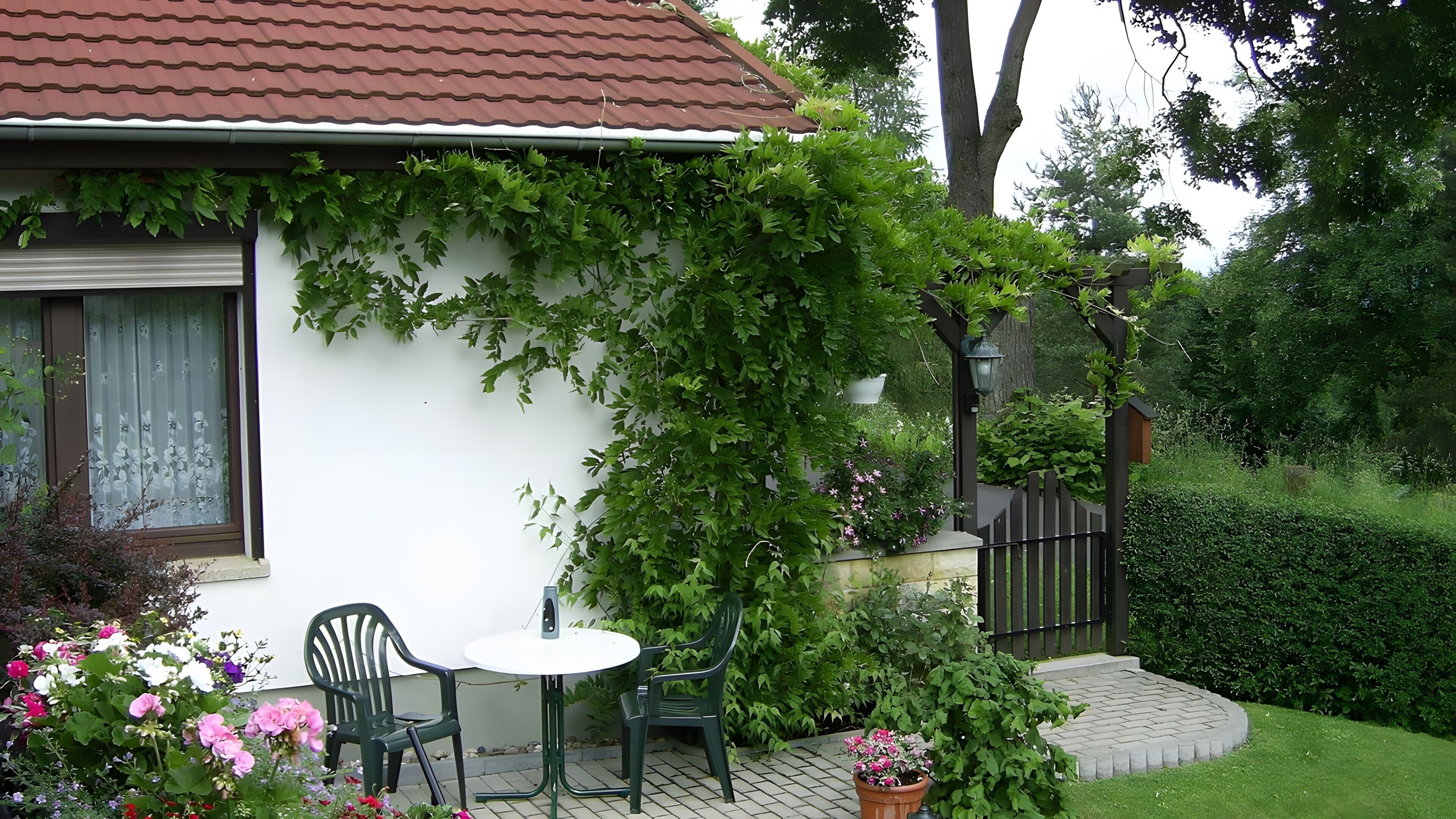 Une terrasse avec une table et des chaises, un bâtiment blanc avec un toit rouge et un jardin où poussent différentes espèces de plantes.
