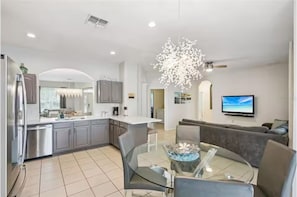 A modern kitchen with grey cabinets, a dining area with a glass table, and a living room with a grey sofa and a TV. - Windsor Hills Lakefront •Walk to Waterpark •2mi WDW•6bd•No Rear Neighbors (Kissimmee)