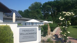 A modern building with a sign welcoming guests, solar panels, and a well-maintained garden with white flowers. - Holiday Apartment 'Meerblick' with Private Terrace, Garden & Wi-Fi (Bastorf)