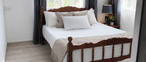 A bedroom with a wooden bed, a bedside table with a lamp, and a view of greenery outside.