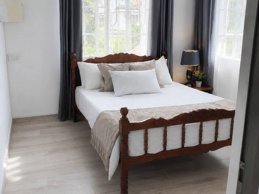 A bedroom with a wooden bed, a bedside table with a lamp, and a view of greenery outside.