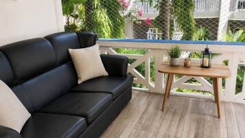 A black leather sofa with a cushion, a small wooden table with a lantern, and a white railing with greenery in the background.