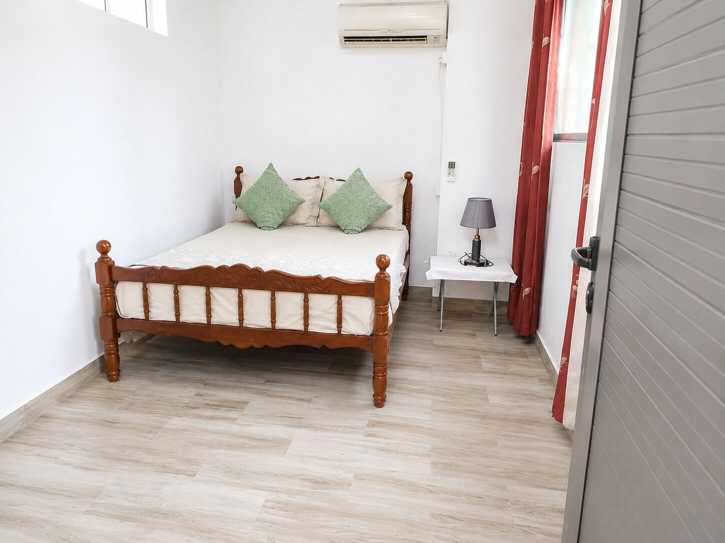 A single bed with a wooden frame and green pillows, a small bedside table with a lamp, and a red curtain.