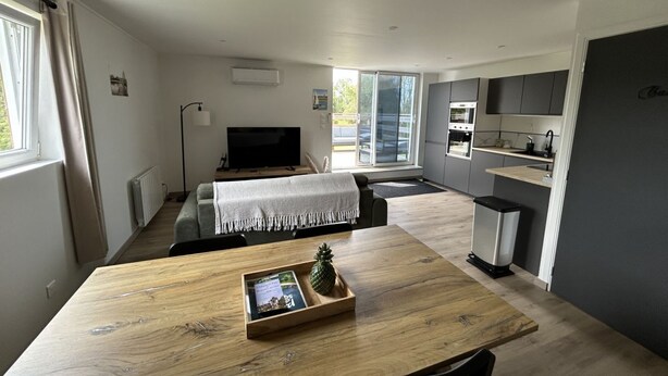 Una cocina moderna con horno integrado, una zona de comedor con mesa de madera y un rincón de estar con sofá y televisor.