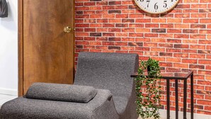 A room with a brick wall, a wooden door, a grey armchair, a small table, and a hanging plant. - Modern 2BR Basement • Near LRT • Parking (Seattle)