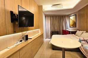 A hotel room with a TV mounted on a wooden panel, a bed, a round table, and a window with curtains. - Shell Residences MOA - The Executive Suites SL005 Cozy 1 Bedroom with AC, Wi-Fi (Pasay City)