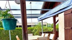 An outdoor patio with a wooden table and chairs, a hanging planter, and a covered area with a view of a building.