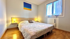 A bedroom with a bed, two bedside lamps, a nightstand, a window with curtains, a radiator, and a painting on the wall.