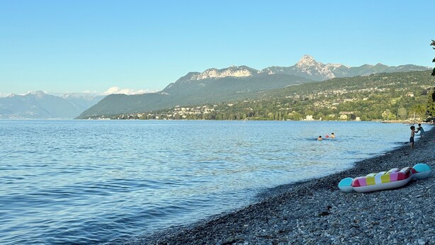 A lake with a pebbled shore, boats, and mountains in the background.