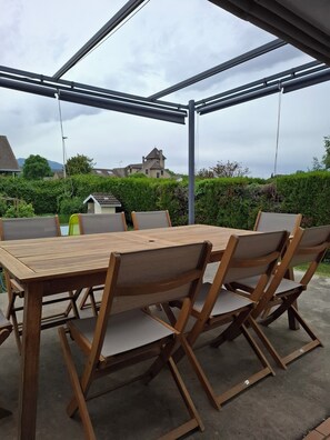 An outdoor patio with a wooden table and chairs, a covered area, and a view of houses and greenery.