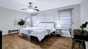 Ein Schlafzimmer mit Bett, Schreibtisch, Stuhl, Deckenventilator und Fenster mit Jalousien.