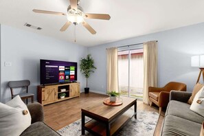 Ein Wohnzimmer mit Flachbildfernseher, Deckenventilator, Couchtisch aus Holz, einer Couch, einem Sessel und einer Topfpflanze.