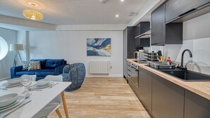 A modern kitchen with a built-in oven, a black sink, and a dining area with a white table and blue sofa. - Alexandra House by Guestz (Stockport)