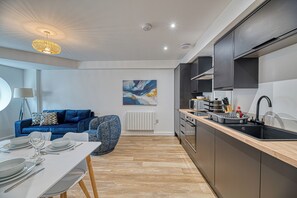 A modern kitchen with a built-in oven, a black sink, and a dining area with a white table and blue sofa. - Alexandra House by Guestz (Stockport)