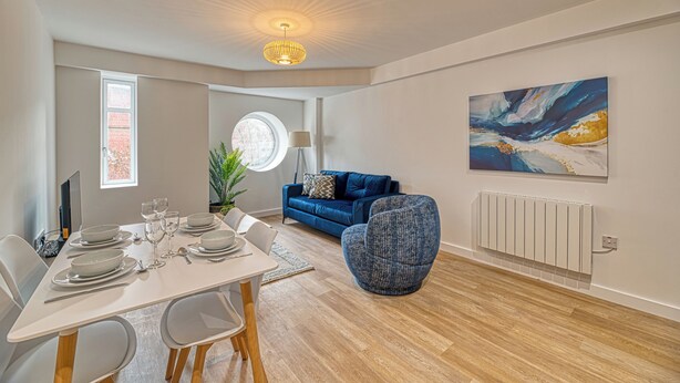 A modern living room with a dining area, a blue sofa, a round window, and a large abstract painting on the wall. - Alexandra House by Guestz (Stockport)