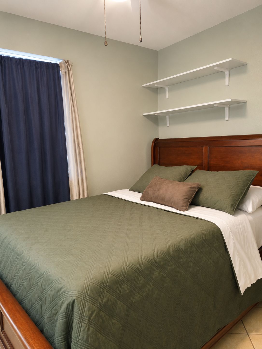 A bed with a wooden headboard, a green bedspread, and two pillows. There are white wall-mounted shelves above the bed.