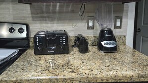 A kitchen with dark wooden cabinets, a stainless steel range hood, a Cuisinart toaster, a blender, and a coffee maker on a granite countertop. - 5BR retreat/Sleeps large groups/Spacious backyard/Near South Padre and Space X. (Brownsville)