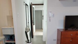A hotel room with a bed, a television on a wooden dresser, a bathroom with a toilet and sink, and a hallway leading to another room.