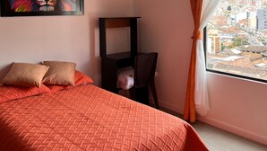 A bedroom with a bed, pillows, a desk, a chair, a painting, and a window with a city view.