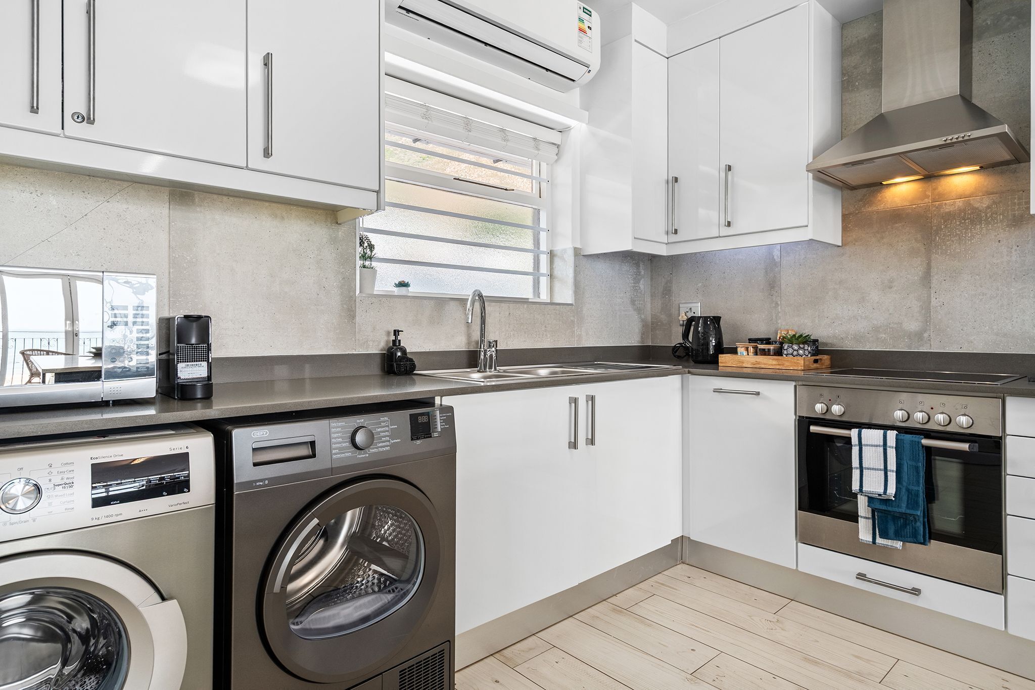 A compact kitchen with a washing machine, a built-in oven, and a window with blinds.