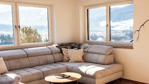 A modern living room with a sectional sofa, a round coffee table, and large windows offering a view of a snowy landscape. - Two beautiful apartments in a sunny and quiet location in Westendorf (Salvenberg)