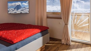 A bedroom with a wooden ceiling, a bed with a red blanket, a window with curtains, and a painting of a mountain. - Two beautiful apartments in a sunny and quiet location in Westendorf (Salvenberg)