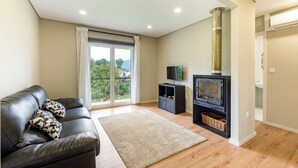 A modern living room with a leather sofa, a flat-screen TV, a fireplace, and a balcony with a view of greenery and buildings.