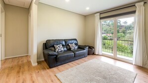 A living room with a black leather sofa, two patterned cushions, a light-colored rug, and a large sliding glass door with a view of greenery.