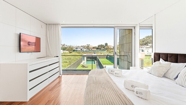 A modern bedroom with a large bed, a TV, and a view of a swimming pool and greenery. - GuestReady - Luxurious Beachfront Villa Retreat (Vila Nova de Gaia)