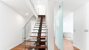A modern staircase with glass railings, leading to a room with a white bed and a light blue accent wall. - GuestReady - Luxurious Beachfront Villa Retreat (Vila Nova de Gaia)