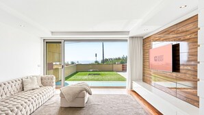 A modern living room with a sofa, a TV mounted on a wooden wall, and a sliding glass door leading to a balcony with a view. - GuestReady - Luxurious Beachfront Villa Retreat (Vila Nova de Gaia)
