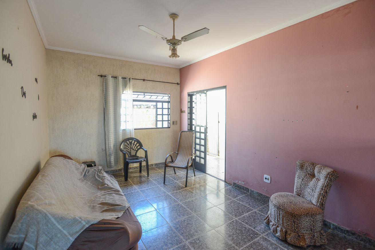 A room with a bed, two chairs, a ceiling fan, a window with curtains, and a door.