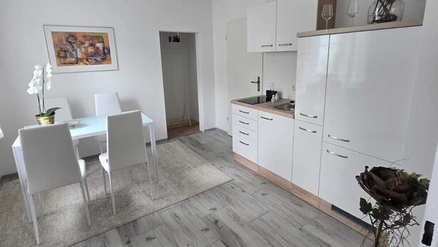 A modern kitchen with white cabinets, a dining area with a white table and chairs, and a grey wooden floor. - Vacation apartment with several living units (up to 20 people) (Wunsiedel)