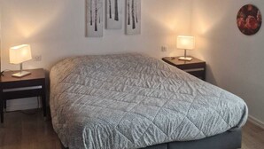 Une chambre à coucher avec un lit, deux tables de chevet avec des lampes, un plafond avec une poutre en bois et trois tableaux accrochés au mur.