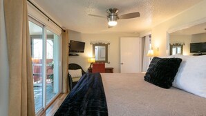 A hotel room with a bed, a ceiling fan, a television, a desk with a lamp, and a sliding glass door leading to an outdoor area.