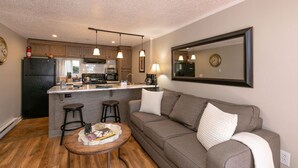 A modern kitchen with a central island, wooden stools, a sofa with cushions, and pendant lighting.