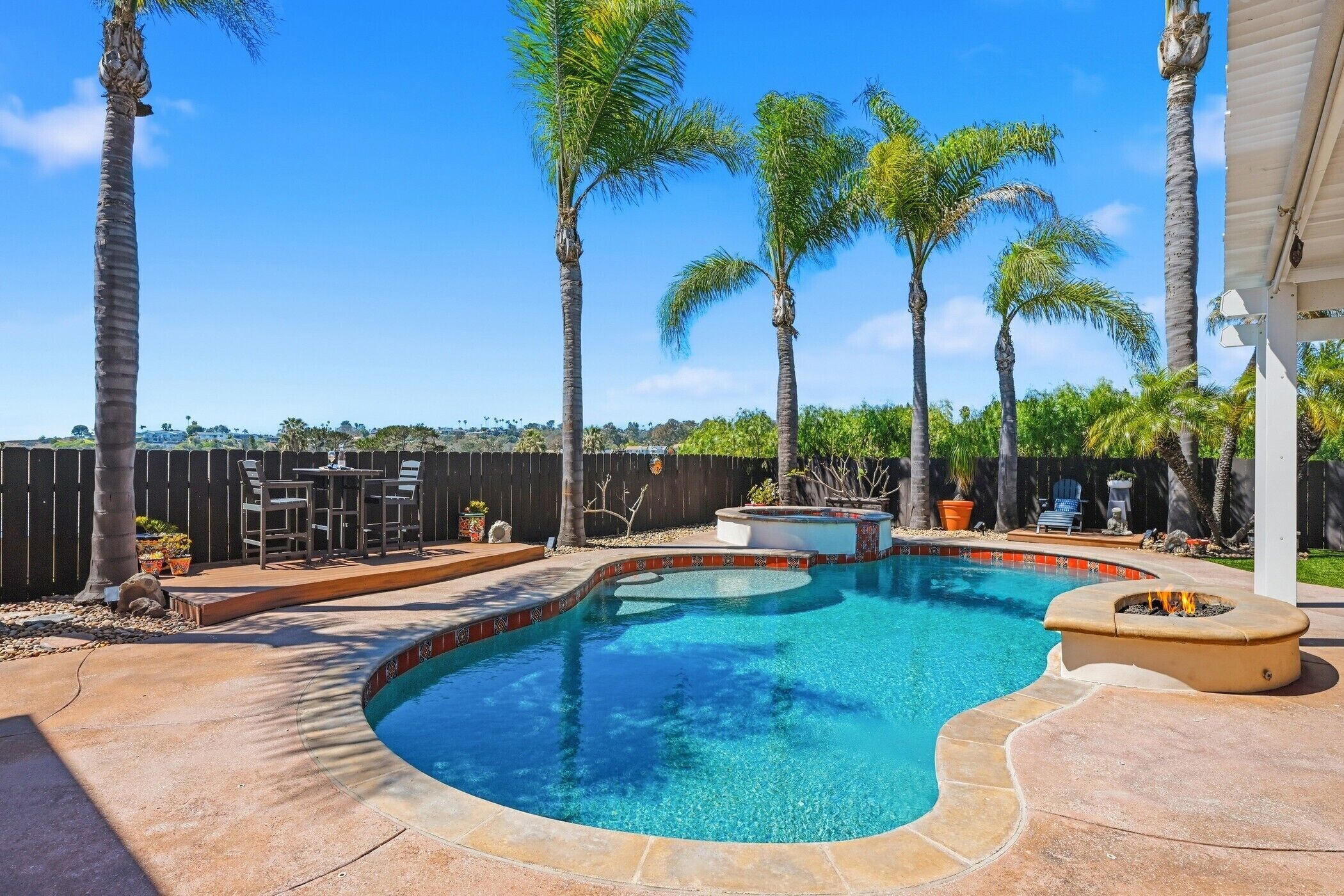 A swimming pool with a curved edge, surrounded by palm trees and a patio area with outdoor furniture.